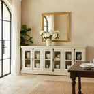 White sideboard with glass doors in a room with a mirror and decorative items.