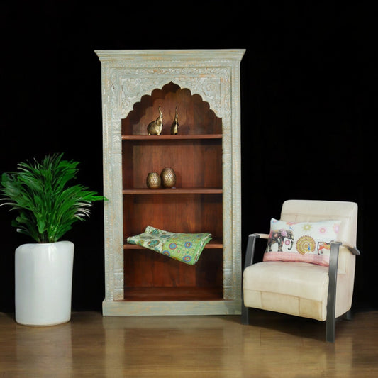 A hand-carved mango wood bookcase with four compartments, placed next to a chair and a potted plant, showcasing its use in a room setting.