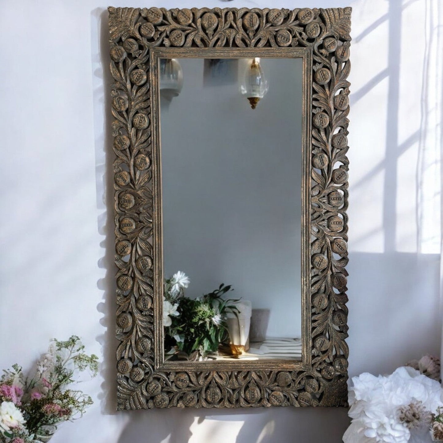 A large, rectangular, handcarved wooden vanity wall mirror with a floral and geometric latticework frame, displayed on a white wall with a mirrored reflection of a garden in the background.