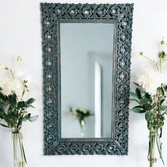 A large, rectangular, handcarved wooden vanity wall mirror with a floral and geometric latticework frame, displayed on a white wall with a mirrored reflection of a garden in the background.